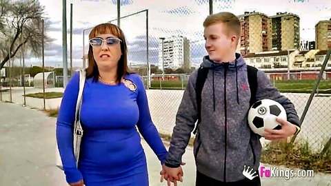 Busty MILF in tight blue dress holds hands with young freckled soccer teen outdoors