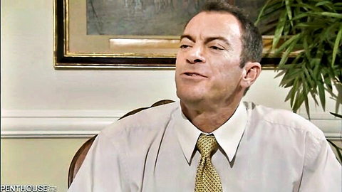 Businessman in white shirt and yellow tie sitting sideways in elegant office with painting and plant.