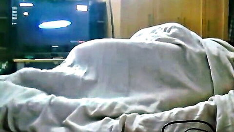 Mysterious bulge under white sheets on bed in dimly lit homemade bedroom with TV glowing.