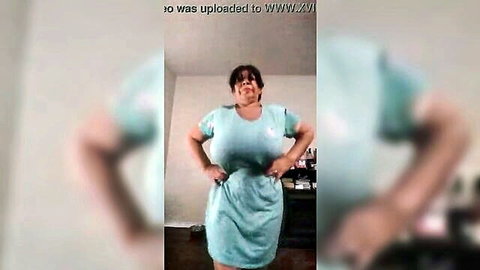 Busty woman in tight turquoise dress posing with hands on hips flaunting massive tits solo