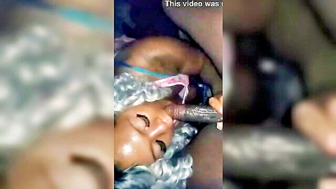 Ebony woman with blue wig lying back, hand holding black dildo to her open mouth