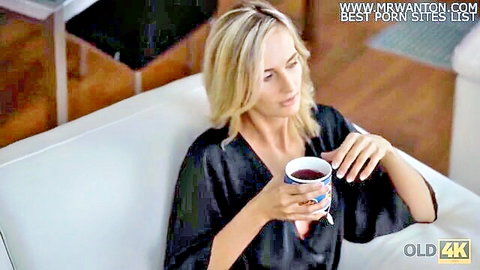 Blonde with small natural tits exposed in open black robe holds mug on couch