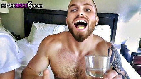 Muscular shirtless bearded hunk grins with open mouth holding glass of water on bed
