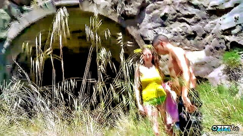 Blonde babe in yellow bikini top and skirt flirts with shirtless muscular hunk outdoors by rocky cave