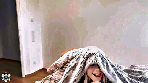 Playful blonde babe peeking out from under gray blanket smiling widely on bed