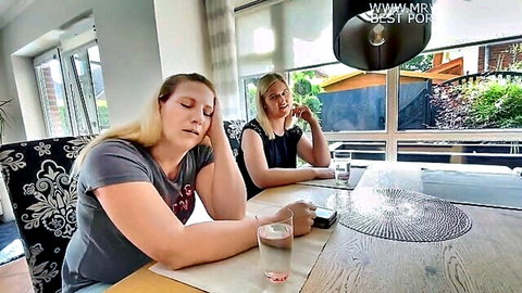 Two blonde Caucasian women sitting clothed at wooden kitchen table chatting casually