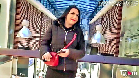 Brunette woman smiling in black puffer jacket holding red purse and phone in public mall walkway