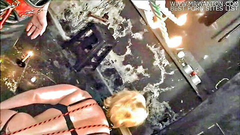Blonde submissive in black strappy harness kneels amid dripping hot wax and lit candles in intense BDSM torture scene