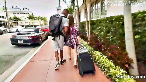 Sara Jay walks with black hubby on sunny sidewalk, suitcase in hand