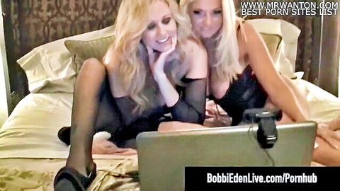 Two busty blondes in sheer black lingerie smiling teasingly on bed with webcam laptop