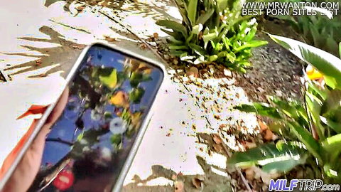 Hand holding smartphone displaying blurred colorful porn thumbnail with MILFTRIP.com and mrwantson.com watermarks outdoors