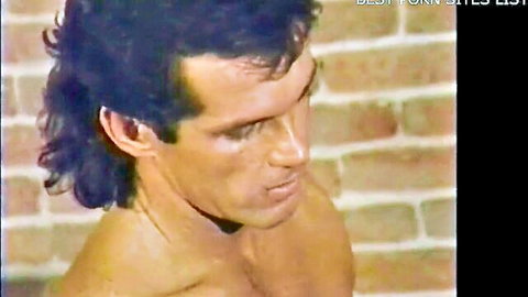 Shirtless muscular hunk with retro mullet hairstyle stares intensely against brick wall
