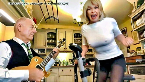 Blonde MILF in sheer blouse pedals exercise bike while older man plays guitar