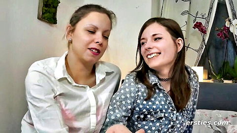 Two smiling amateur Caucasian girls in blouses sitting close on bed teasing for lesbian fun