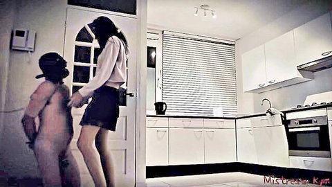 Brunette femdom mistress towers over naked hooded submissive slave in kitchen BDSM domination