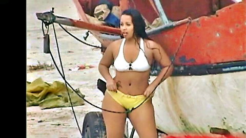 Curvy Latina in yellow bikini pulls down bottoms on beach near boat, teasing public exposure