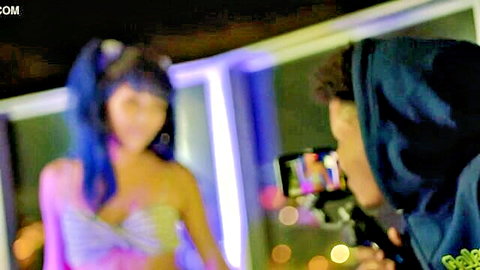 Topless young blue-haired Asian teen with small tits exposes nipples while hooded guy films her seductively.