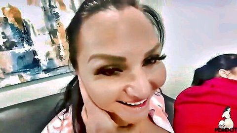 Busty smiling Latina in pink low-cut top touching neck with curvy friend in red hoodie on couch