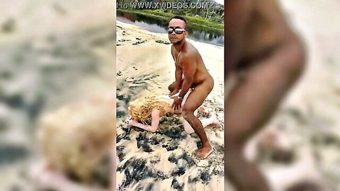 Nude black man in sunglasses stands over blonde woman in yellow dress on beach sand