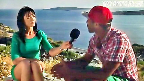 Brunette MILF reporter in green dress interviews black stud seaside holding mic with legs spread teasing