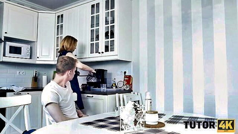 Blonde MILF tutor in kitchen pours hot drink for seated young man student