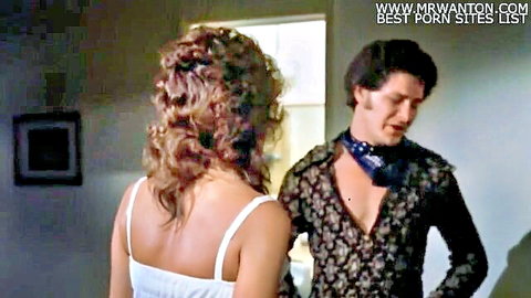 Vintage blonde in tight white dress stands close to man in floral shirt and scarf in retro bedroom scene