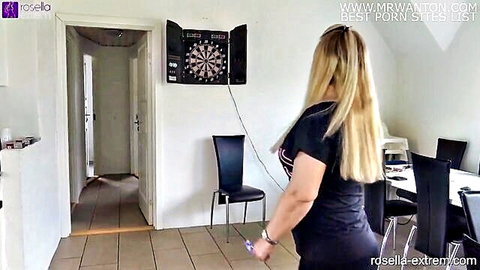 Blonde MILF in tight black top and long hair walking toward dartboard in kitchen room