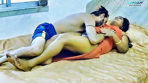 Muscular shirtless Indian man passionately kisses and hugs busty mature woman in hiked orange dress exposing thighs on bed