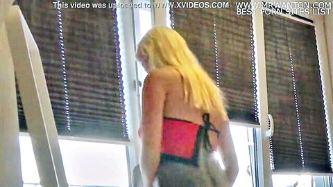 Blonde teen in red backless top stands by window at house party by Leon Lambert