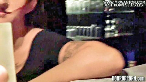 Tattooed woman in black top leans over bar counter with red lips pursed seductively