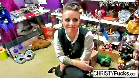 Tattooed fair-skinned brunette with mohawk squats smiling in cluttered nerdy room full of toys