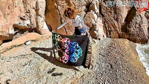 Brunette babe in tiny shorts holding colorful skull BAY BAY blanket on rocky beach outdoor tease