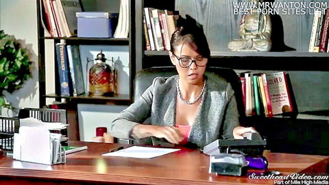 Sexy brunette MILF in glasses and gray blazer sitting at office desk with papers