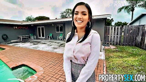 Smiling Latina real estate agent in blouse and skirt poses by backyard pool
