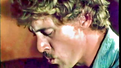 Close-up of vintage blonde curly-haired man with mustache smirking in retro adult video