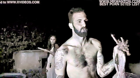 Shirtless tattooed bearded hunk smokes cigarette holding raw meat leg by foggy outdoor bbq pit with woman watching