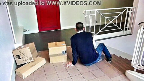 Man in sweater sitting on tiled floor surrounded by cardboard boxes in modern hallway with red door
