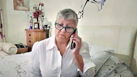 Mature busty woman in white blouse talks on phone in bedroom