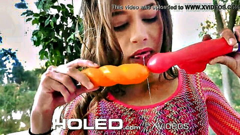 Brunette babe sucking red dildo with saliva dripping while gripping orange toy outdoors