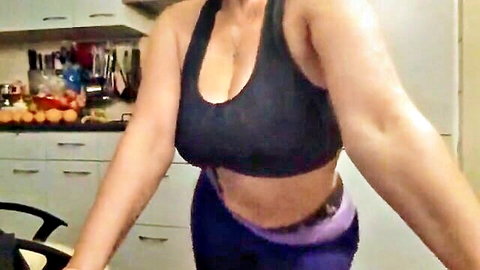 Busty muscular woman in tight black sports bra flaunts massive cleavage and toned arms in kitchen wearing purple leggings