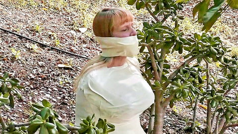 Blonde woman tightly wrapped in white microfoam tape, gagged and bound in outdoor BDSM mummy fetish scene