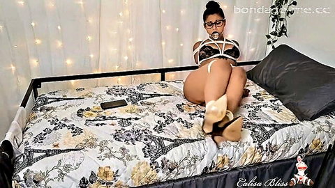Bound brunette in ball gag BDSM harness spreads thick thighs teasing pussy on bed