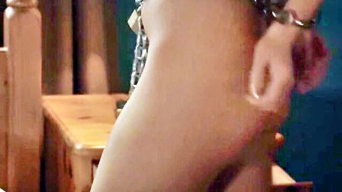Brunette submissive in heavy BDSM chains exposing naked tits and curves beside wooden bed