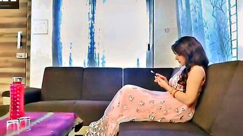 Brunette South Asian beauty in pink floral dress sits legs crossed on couch scrolling phone