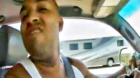 Black man with mustache intensely smiling while driving car in white tank top