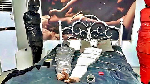 Woman mummified in white duct tape and saran wrap bound spread-eagle on bed by latex-clad figures