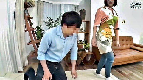 Japanese woman in colorful apron stands behind kneeling man in domestic JAV living room scene