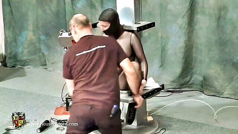 BDSM setup with hooded submissive restrained in device by bald dominant