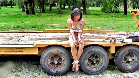 Brunette in sexy bra with tits spilling out, wrists and ankles bound in outdoor bondage on yellow trailer