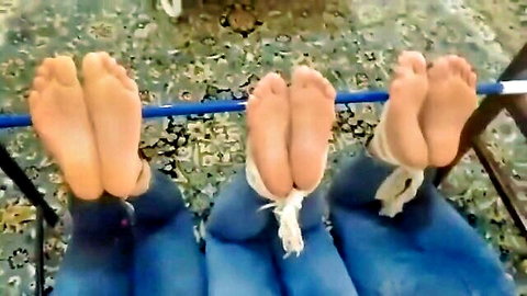 Three women's bare soles bound with ropes to blue bar in BDSM foot worship pose